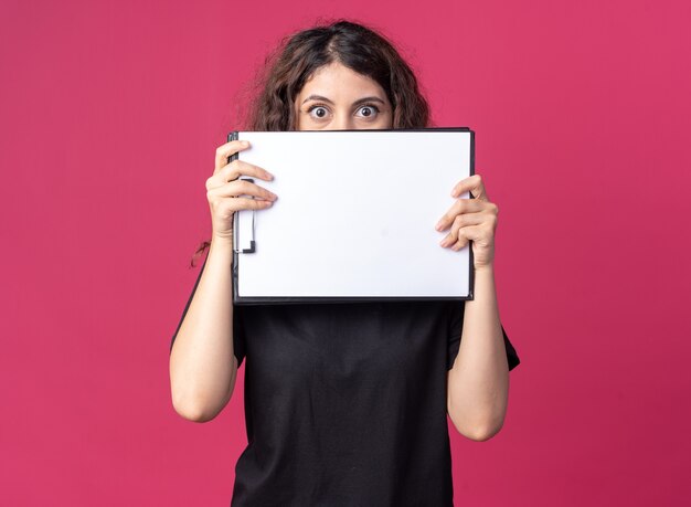 Impressed young pretty girl holding clipboard from behind it 