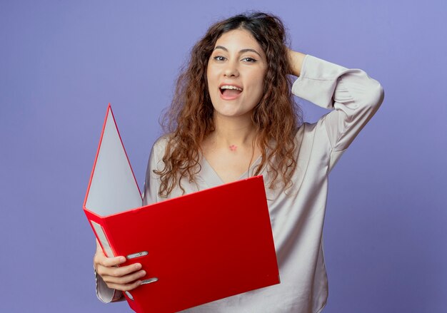 Impressed young pretty girl holding folder and putting hand behind head 