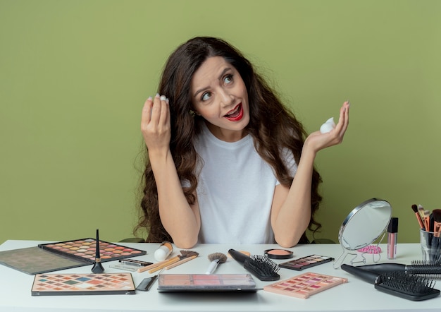 Impressionato giovane bella ragazza seduta al tavolo per il trucco con strumenti di trucco che applica la mousse per capelli sui capelli e guardando il lato isolato su sfondo verde oliva