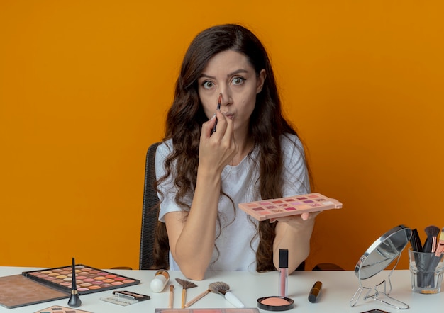 Free photo impressed young pretty girl sitting at makeup table with makeup tools holding eyeshadow palette and touching nose with eyeshadow brush isolated on orange background