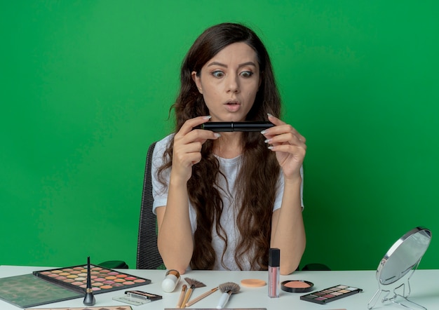 Impressed young pretty girl sitting at makeup table with makeup tools holding and looking at mascara isolated on green background