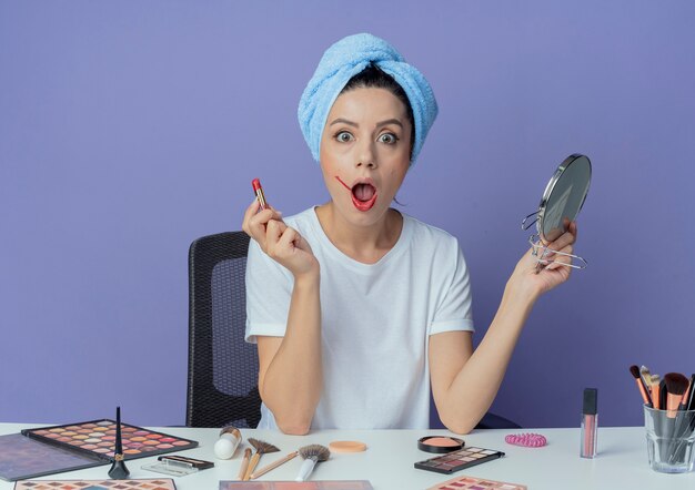 Impressed young pretty girl sitting at makeup table with makeup tools and with bath towel on head holding mirror and holding red lipstick isolated on purple background