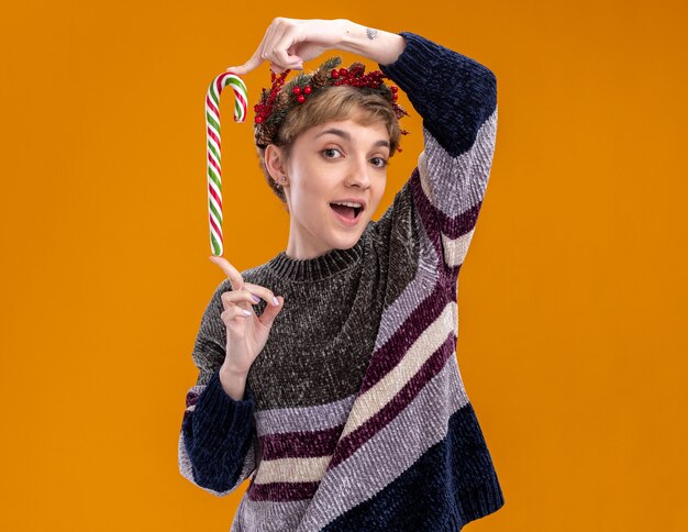 Impressed young pretty girl wearing christmas head wreath holding christmas sweet cane looking at camera isolated on orange background