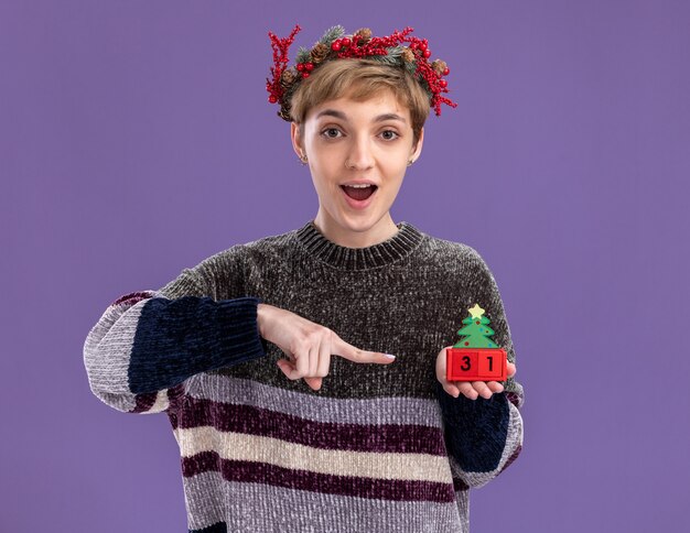 Impressed young pretty girl wearing christmas head wreath holding christmas tree toy with date pointing at it  isolated on purple wall