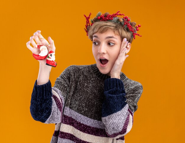 Impressed young pretty girl wearing christmas head wreath holding and looking at santa claus christmas ornaments keeping hand on face isolated on orange wall with copy space