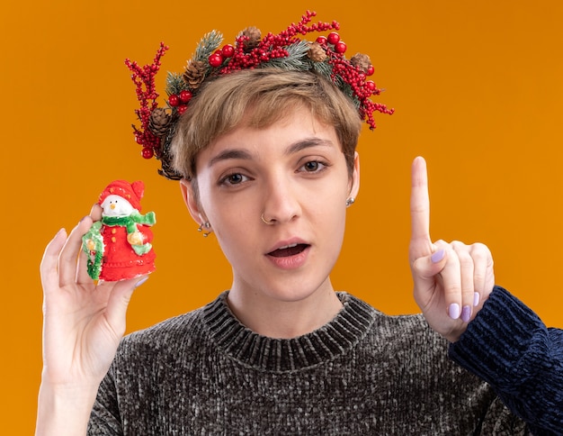 Impressed young pretty girl wearing christmas head wreath holding small christmas snowman statue  pointing up isolated on orange wall