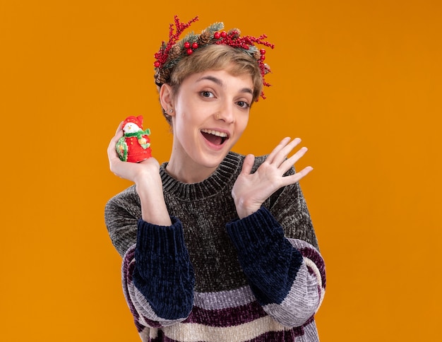 コピースペースでオレンジ色の壁に分離された空の手を示す小さなクリスマス雪だるま像を保持しているクリスマスの頭の花輪を身に着けている感動の若いかわいい女の子