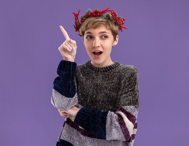 Impressed young pretty girl wearing christmas head wreath looking at side pointing up isolated on purple background