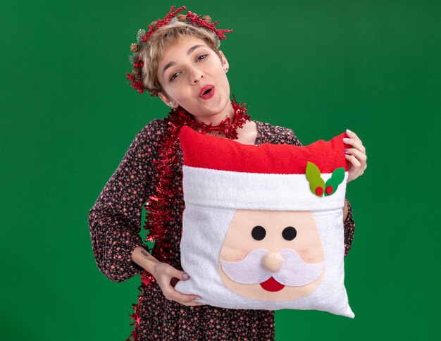 Impressed young pretty girl wearing christmas head wreath and tinsel garland around neck holding santa claus pillow  isolated on green wall