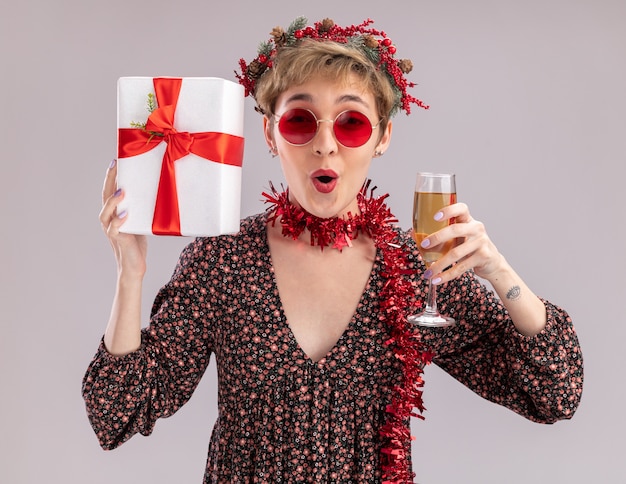 Impressed young pretty girl wearing christmas head wreath and tinsel garland around neck with glasses holding gift package and glass of champagne looking at camera isolated on white background