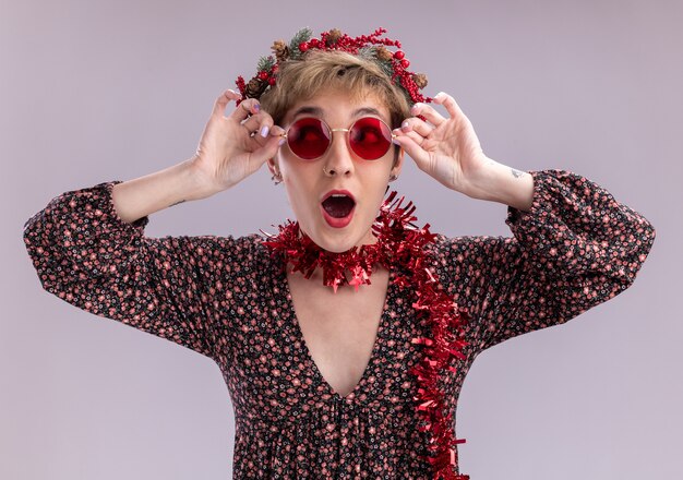 Impressed young pretty girl wearing christmas head wreath and tinsel garland around neck with glasses looking at side grabbing glasses isolated on white wall