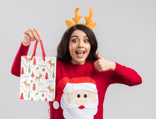 Impressed young pretty girl wearing reindeer antlers headband and santa claus sweater holding christmas gift bag looking showing thumb up 