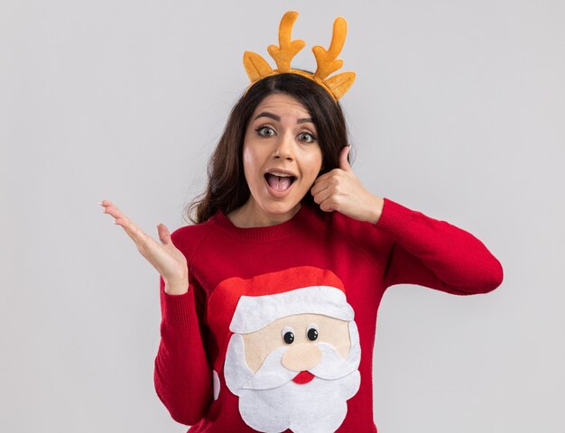 Impressed young pretty girl wearing reindeer antlers headband and santa claus sweater looking showing empty hand and thumb up 