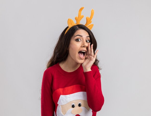 Impressed young pretty girl wearing reindeer antlers headband and santa claus sweater looking whispering 