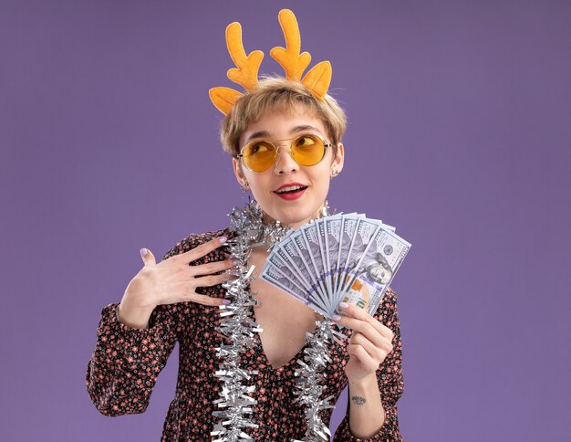 Impressed young pretty girl wearing reindeer antlers headband and tinsel garland around neck with glasses holding money looking up touching shoulder isolated on purple wall with copy space