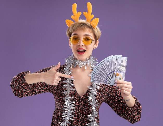 Impressed young pretty girl wearing reindeer antlers headband and tinsel garland around neck with glasses holding and pointing at money looking at camera isolated on purple background