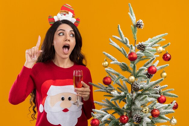 впечатленная молодая красивая девушка в повязке на голову санта-клауса и свитере, стоящая возле украшенной рождественской елки с бокалом шампанского, смотрящая в сторону, указывающую вверх, изолирована на оранжевом фоне