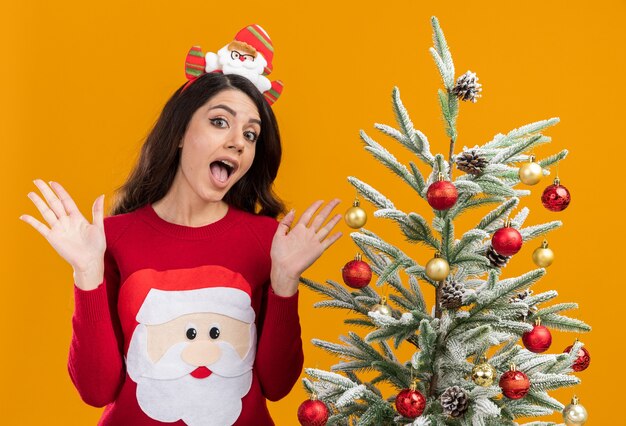 オレンジ色の背景で隔離の空の手を示すカメラを見て装飾されたクリスマスツリーの近くに立っているサンタクロースのヘッドバンドとセーターを着ている感動の若いかわいい女の子