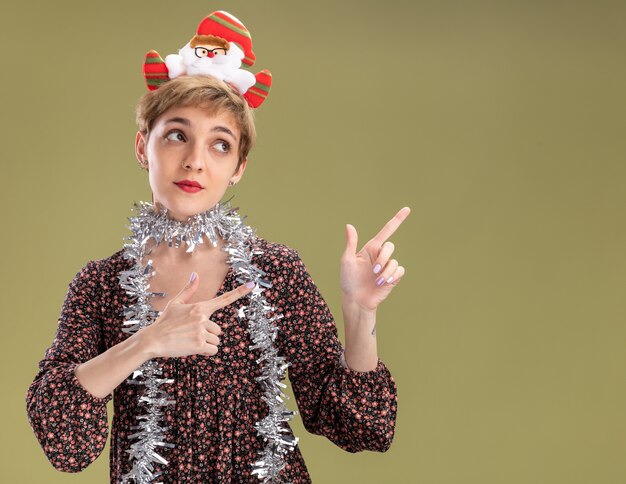 Impressionato giovane bella ragazza che indossa la fascia di babbo natale e la ghirlanda di orpelli intorno al collo guardando e indicando il lato isolato sulla parete verde oliva con lo spazio della copia