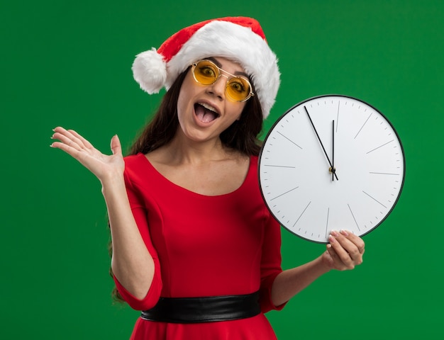 Impressionata giovane ragazza carina che indossa cappello da babbo natale e occhiali che tengono l'orologio che mostra la mano vuota isolata sul muro verde