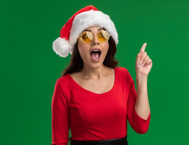 Free photo impressed young pretty girl wearing santa hat and glasses looking at side pointing up isolated on green background