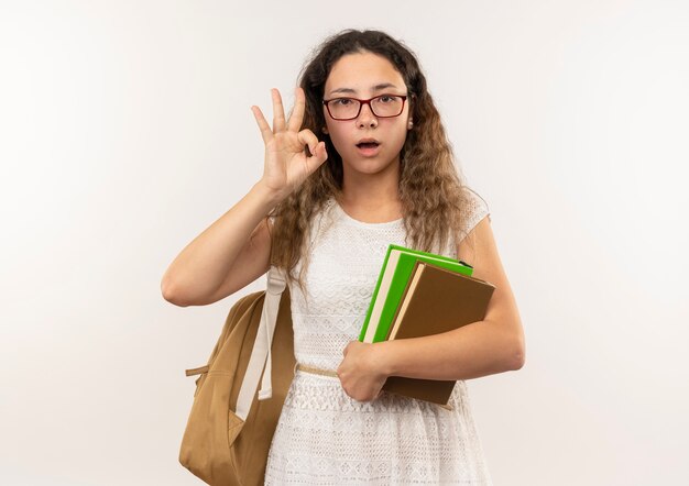 Impressed young pretty schoolgirl wearing glasses and back bag holding books doing ok sign isolated on white