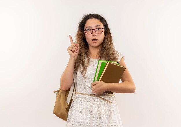 Впечатленная молодая симпатичная школьница в очках и задней сумке, держащая книги, поднимая палец на белом