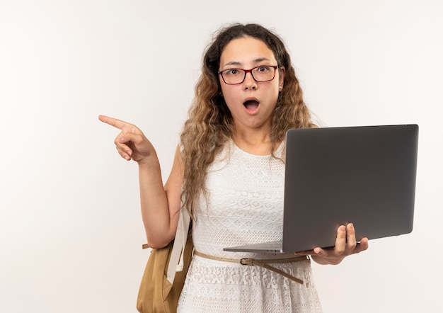 Impressed young pretty schoolgirl wearing glasses and back bag holding laptop and pointing at side isolated on white