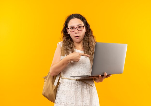 안경을 착용하고 다시 가방을 들고 노란색에 고립 된 노트북을 가리키는 감동 젊은 예쁜 여학생