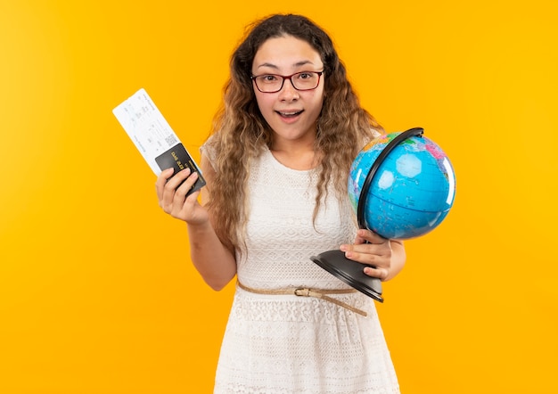 Impressionato giovane studentessa graziosa con gli occhiali in possesso di biglietti aerei, carta di credito e globo isolato su giallo
