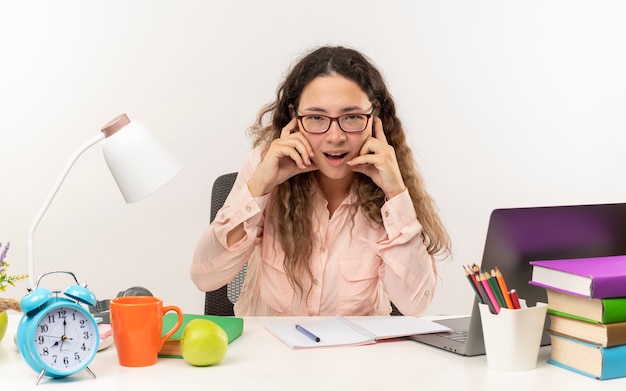 Impressed young pretty schoolgirl wearing glasses sitting at desk with school tools doing her homework putting fingers on ears isolated on white