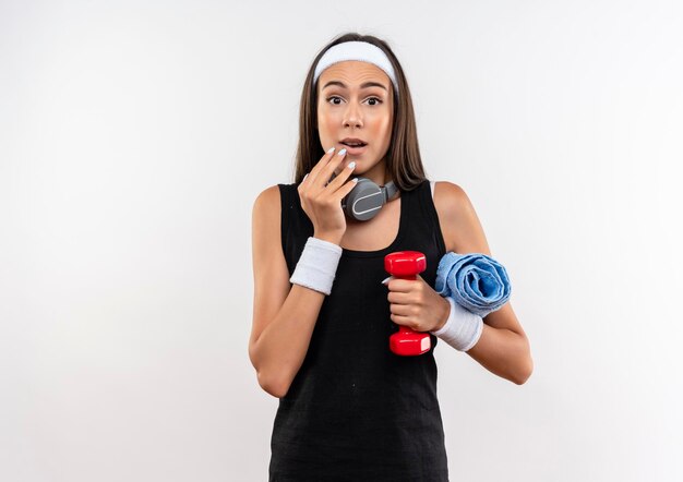 Impressed young pretty sporty girl wearing headband and wristband and headphones on neck holding dumbbell with towel putting hand on chin isolated on white wall with copy space