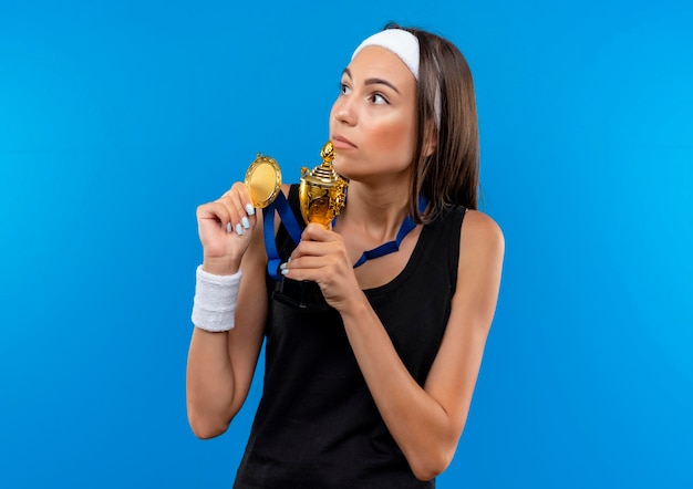 그녀의 목에 머리띠와 팔찌와 메달을 착용하고 우승 컵과 메달을 들고 파란색 벽에 고립 된 측면을보고 감동 젊은 꽤 스포티 한 소녀