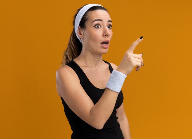 Free photo impressed young pretty sporty girl wearing headband and wristbands looking and pointing at side isolated on orange wall with copy space