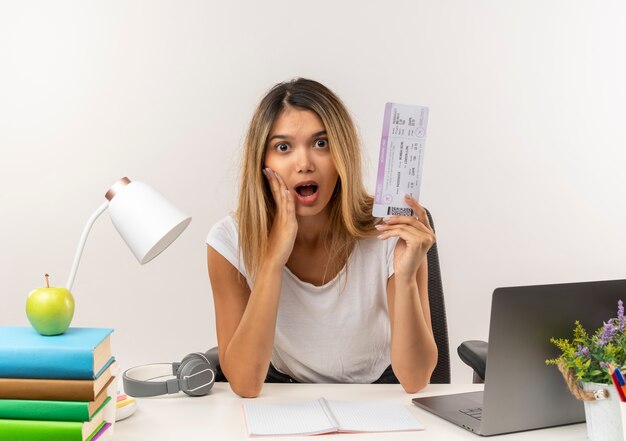 Impressed young pretty student girl sitting at desk with school tools putting hand on cheek and holding airplane ticket isolated on white