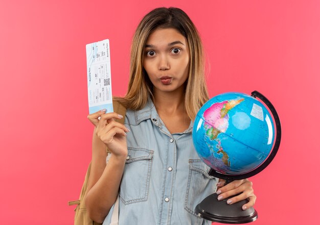 Impressed young pretty student girl wearing back bag holding airplane ticket and globe isolated on pink
