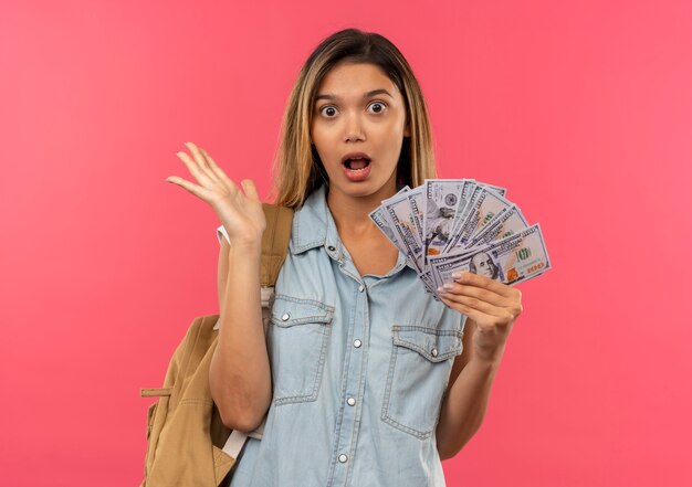 Impressed young pretty student girl wearing back bag holding money and showing empty hand isolated on pink