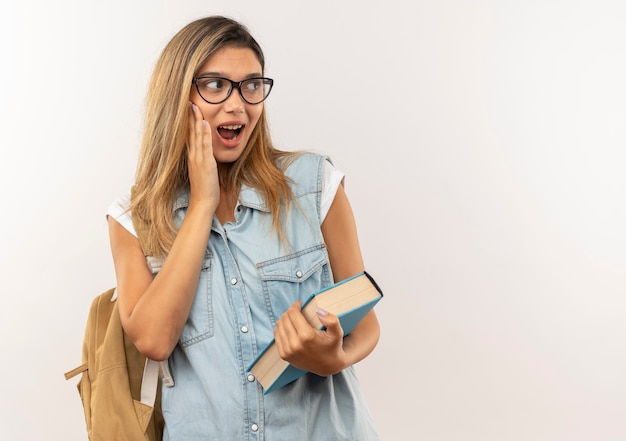 Impressionato giovane ragazza graziosa studentessa con gli occhiali e borsa posteriore che tiene il libro loooking a lato con la mano sulla guancia isolato su bianco