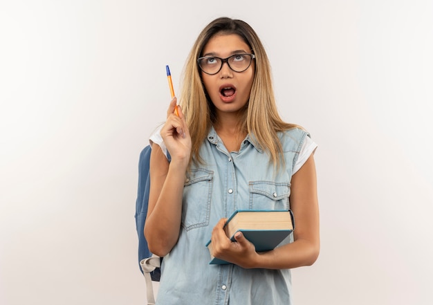 Impressed young pretty student girl wearing glasses and back bag holding book and pen looking up isolated on white