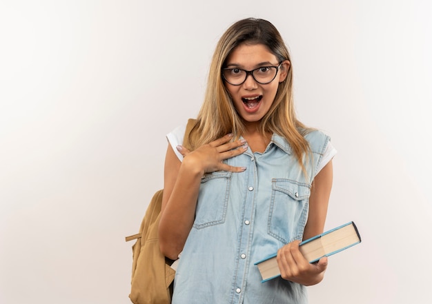 Impressionato giovane ragazza graziosa studentessa con gli occhiali e borsa posteriore che tiene il libro con la mano sulla guancia isolato su bianco