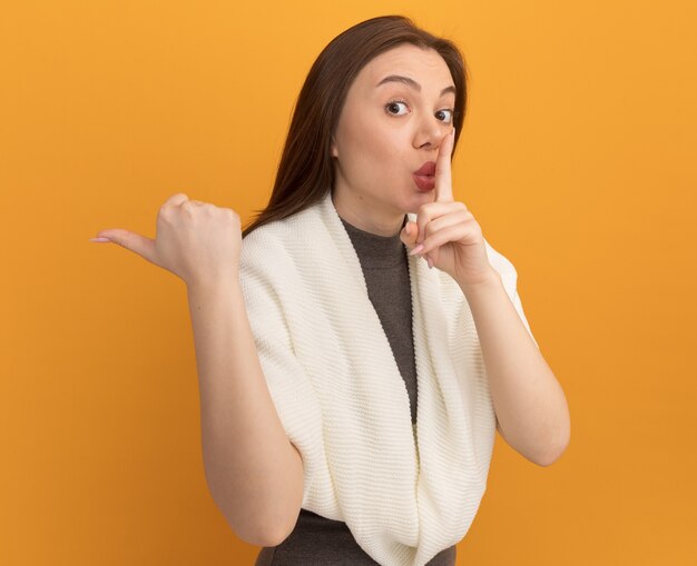 Impressed young pretty woman  doing silence gesture pointing at side isolated on orange wall