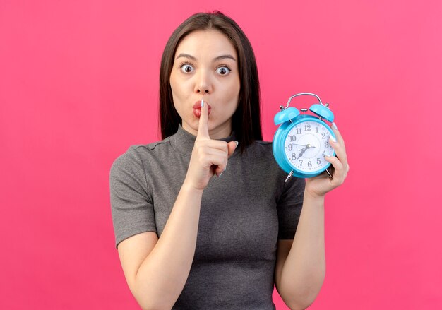 Impressed young pretty woman holding alarm clock and gesturing silence isolated on pink background with copy space