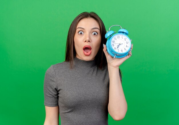 impressed young pretty woman holding alarm clock looking at front