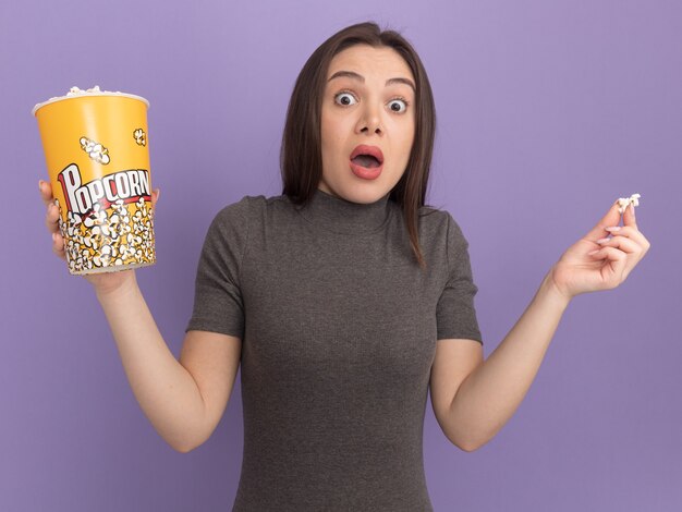 Впечатленная молодая красивая женщина, держащая ведро попкорна и кусок попкорна, изолированные на фиолетовой стене