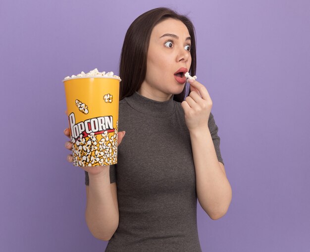 Impressed young pretty woman holding bucket of popcorn and popcorn piece near mouth looking at side 