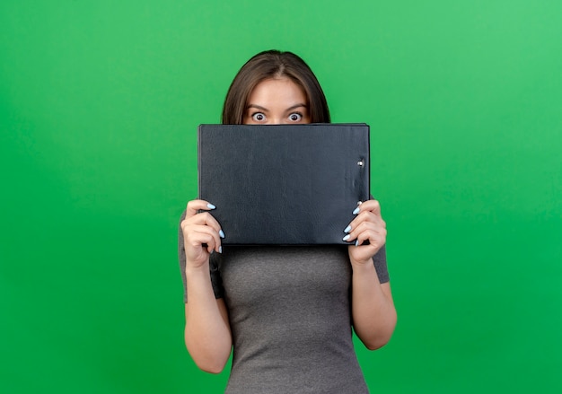 Impressed young pretty woman holding clipboard and looking at camera from behind it isolated on green background with copy space