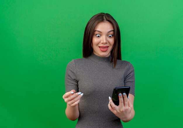 Впечатленная молодая красивая женщина, держащая и смотрящая на мобильный телефон и держащая руку в воздухе, изолированную на зеленом фоне с копией пространства
