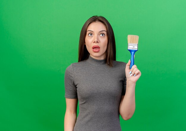 Impressed young pretty woman holding paint brush looking up isolated on green background with copy space