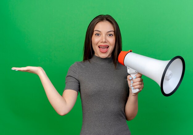 Impressed young pretty woman holding speaker and showing empty hand isolated on green background