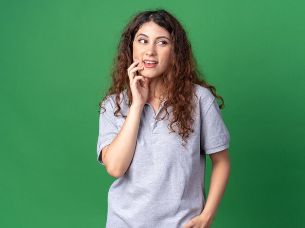 Impressed young pretty woman touching chin looking at side isolated on green wall with copy space